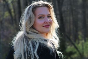 Blonde woman with wavy hair smiling outdoors in natural lighting against a dark forest background.