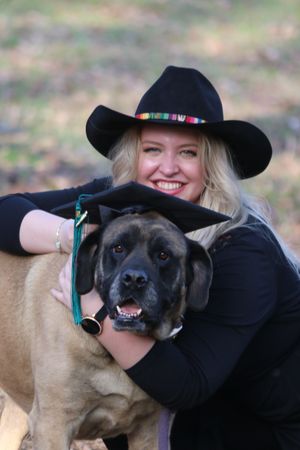 A large brown mixed-breed dog poses with someone wearing a black cowboy hat and embracing the pet outdoors.