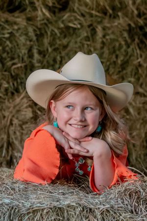 A child wearing a western cowboy hat and orange shirt poses on hay bales with turquoise earrings.