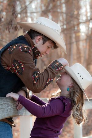 Two people wearing white cowboy hats share a tender moment by a wooden fence in an autumn forest setting.