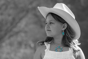 Young girl in white cowboy hat and dress with turquoise jewelry gazes into the distance in selective color photo.
