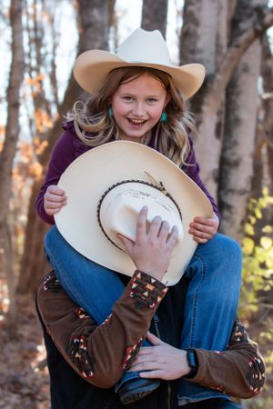 Two cowgirls in western attire share a playful moment with cowboy hats in a forest setting.
