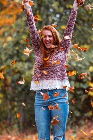 Someone wearing a sweater and jeans joyfully throws autumn leaves in the air during fall season in a park setting.
