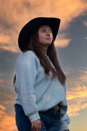 A western-style silhouette at sunset wearing a black hat, white sweater and jeans against dramatic orange clouds.