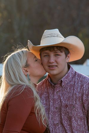 A mother and son sharing a tender moment outdoors, with the mom kissing the cowboy-hat wearing man in a paisley shirt.