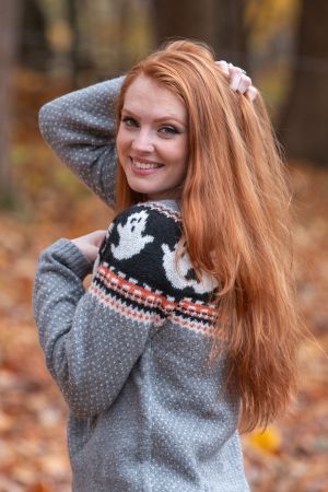 Cozy autumn portrait of long red hair in a gray patterned sweater with fall foliage in the background.