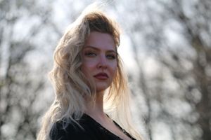 A woman with windswept blonde hair poses outdoors against a blurred natural background at dusk.