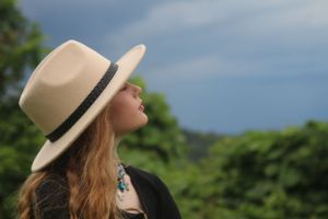 Side profile of someone in a beige panama hat looking upward against green foliage and blue sky outdoors.