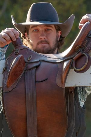 A western-style leather saddle with rich brown leather and decorative details being held up by someone in a cowboy hat.