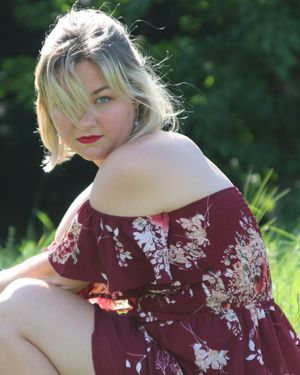 Person in burgundy floral off-shoulder dress poses outdoors in natural sunlight against green foliage.