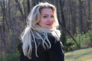 A portrait of someone with long blonde wavy hair wearing a black jacket outdoors in front of a wooded background.
