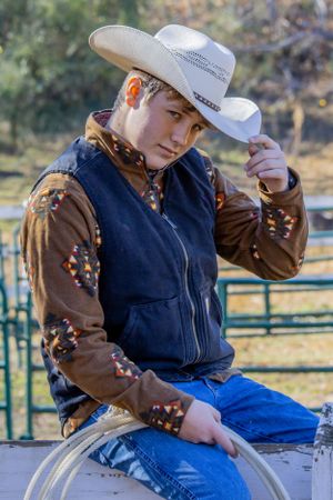 A person in western wear with blue jeans, navy vest, brown patterned jacket and white cowboy hat sits on a fence post.