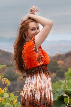 Person in vibrant orange dress poses elegantly among grapevines with mountains in background on cloudy day.