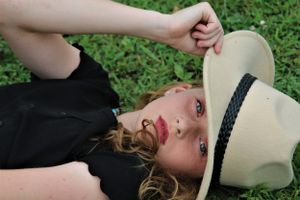 Person lying in grass with a white sun hat, wearing a black outfit against a green natural background.