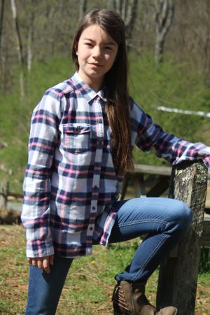 Person in plaid flannel shirt and jeans poses casually by wooden fence in wooded setting.