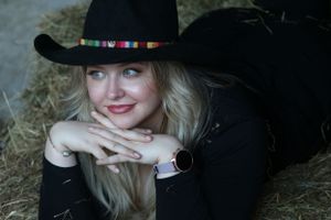 A woman wearing a black cowboy hat and black top rests her chin on folded hands while posing against a hay backdrop.