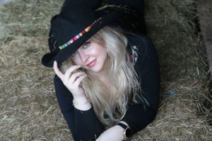 A person wearing a black hat and dark clothing lies in hay while holding the brim of their hat with one hand.