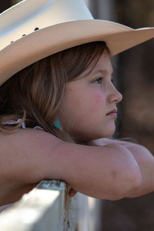 Profile view of a young cowgirl wearing a tan hat and turquoise earrings leaning on a fence in soft lighting.