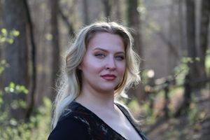 Portrait of someone with blonde hair and a black top posing in a sunny wooded forest setting.