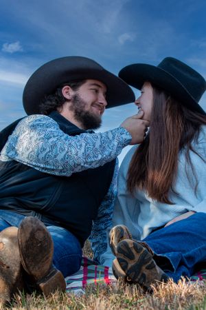 Two people in cowboy hats sit close together on the grass against a blue sky background.