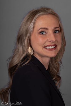 Professional studio portrait against a dark grey background featuring business attire and gold earrings.