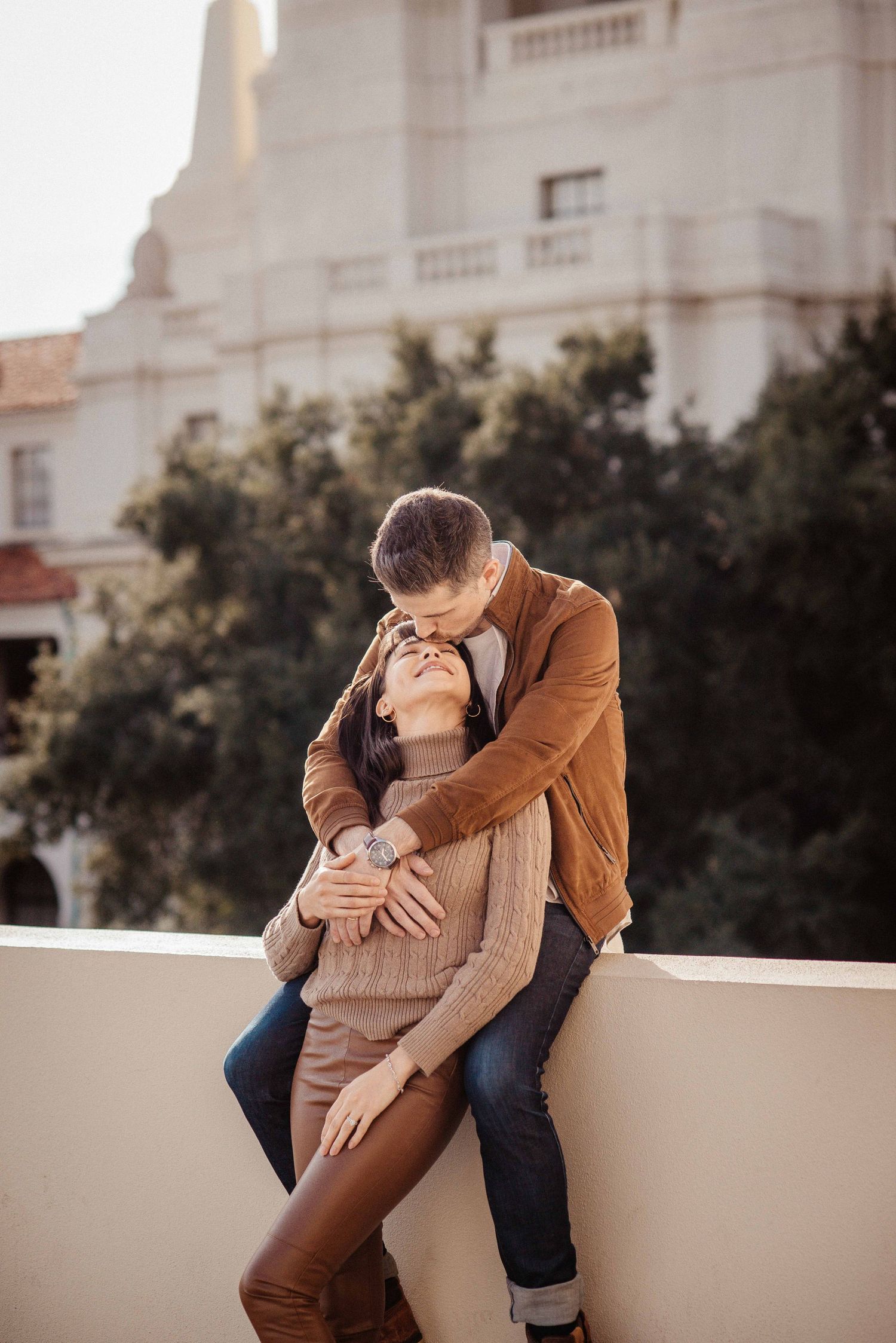 A couple shares an intimate moment on a white stone wall with classic architecture in the background.