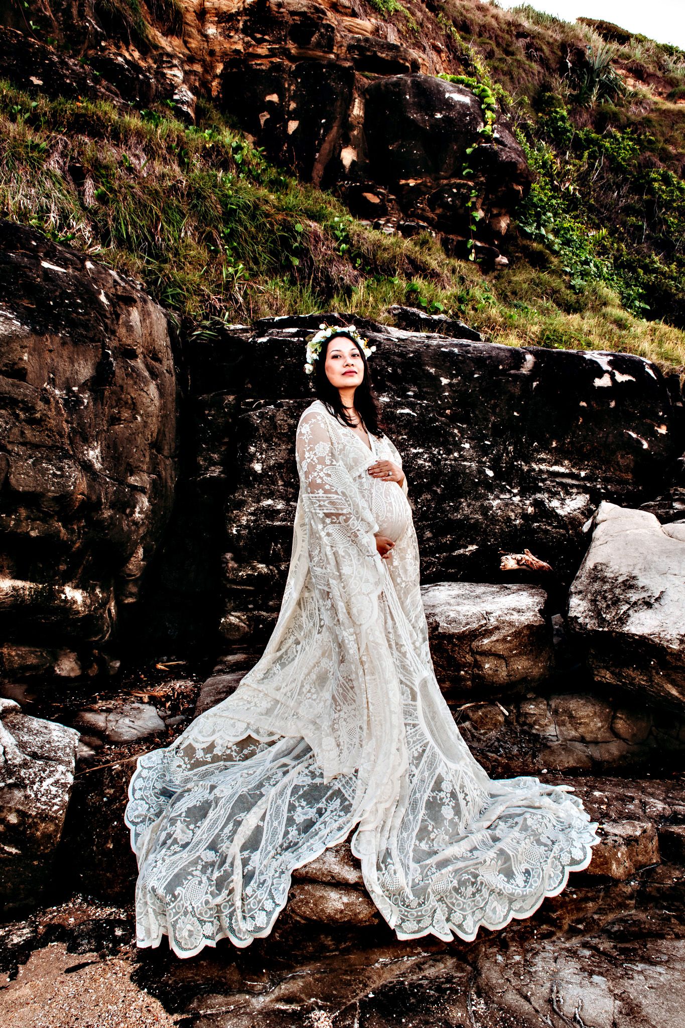 A white lace dress flows dramatically against rocky coastal cliffs.