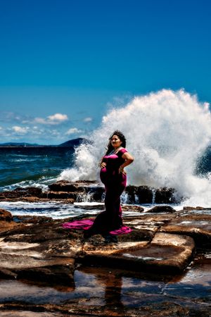 Dramatic ocean waves crash against rocky coastline as figure in purple dress poses on rocks.