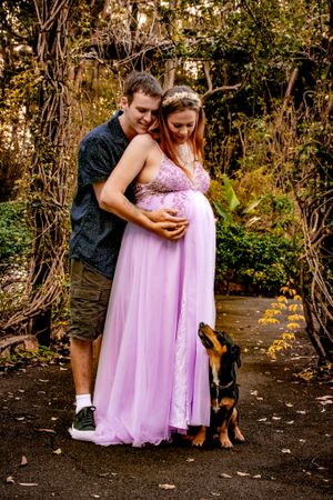 Couple and dog pose together in garden setting with purple maternity gown.