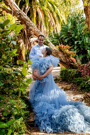 Elegant maternity session featuring a flowing blue ruffled gown in a tropical garden setting.