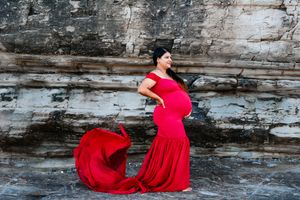 Dramatic maternity photo with flowing red dress against stone cliff backdrop.