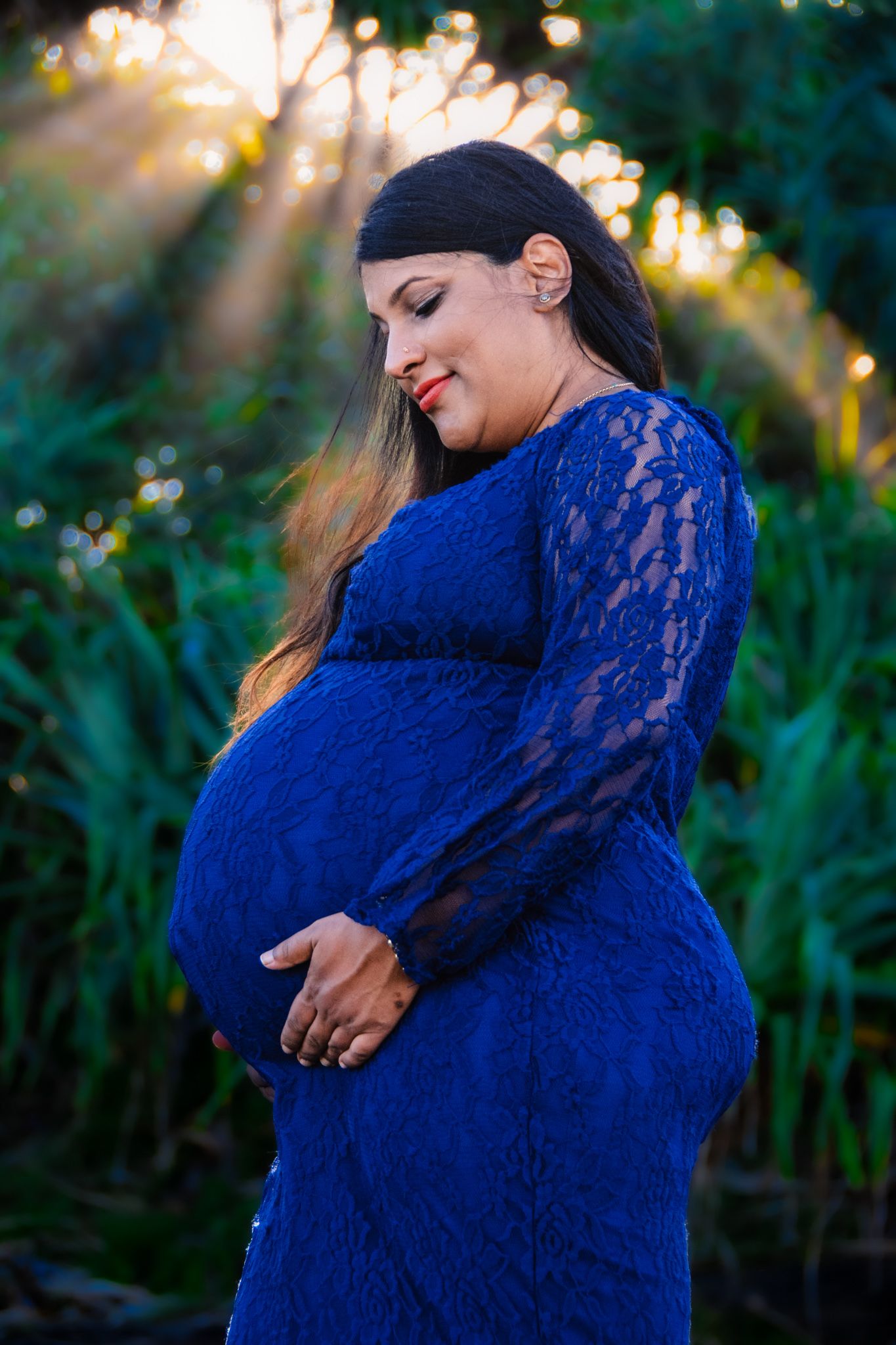 Maternity portrait in blue lace dress with sunlight streaming through trees.