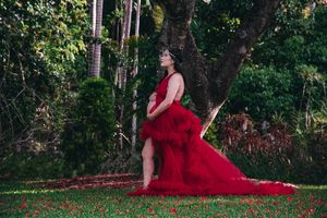 Elegant maternity portraits in flowing red gown surrounded by lush garden scenery.