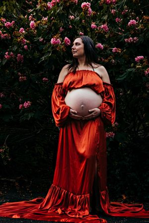 Vibrant red maternity gown contrasts with pink flowering bushes outdoors.