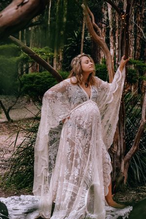 Expectant mother in lace dress poses dramatically among trees in ethereal forest scene.