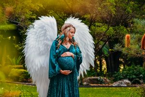 Maternity photo featuring woman in teal dress with white angel wings in garden setting.