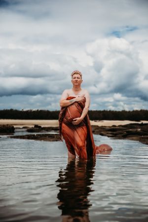 Artistic water portrait session in rustic copper-toned dress against cloudy landscape.