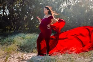 Woman in a flowing red dress poses dramatically against a natural outdoor backdrop.