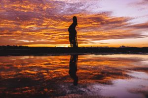 Silhouetted figure against vibrant orange and purple sunset sky over water.