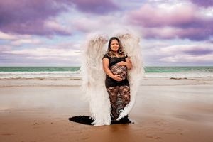 Beach maternity photography with angel wings against purple sunset sky.