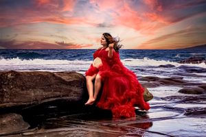 Red gown flows dramatically over rocky coastline during vibrant sunset.