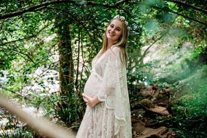 Ethereal maternity photoshoot in a sunlit forest with flowing white lace dress and natural greenery.
