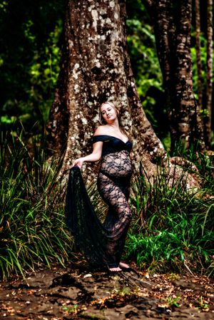 Artistic maternity photos against tree trunks with black lace dress in forest setting.