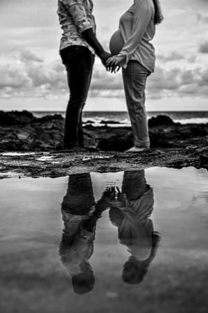 Black and white reflection of couple's feet in tidal pool on rocky beach.