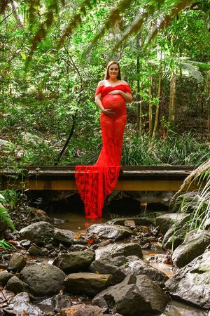 Woman in flowing red dress poses on rocky terrain surrounded by lush forest.