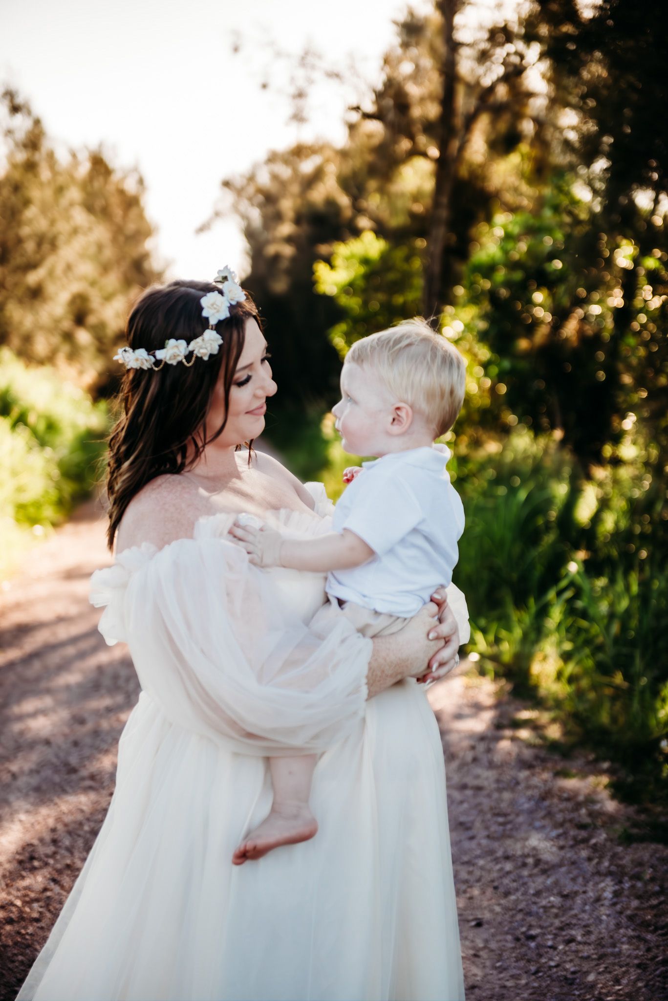 A mother and child share tender moments in flowing white dresses against a sunlit forest path.