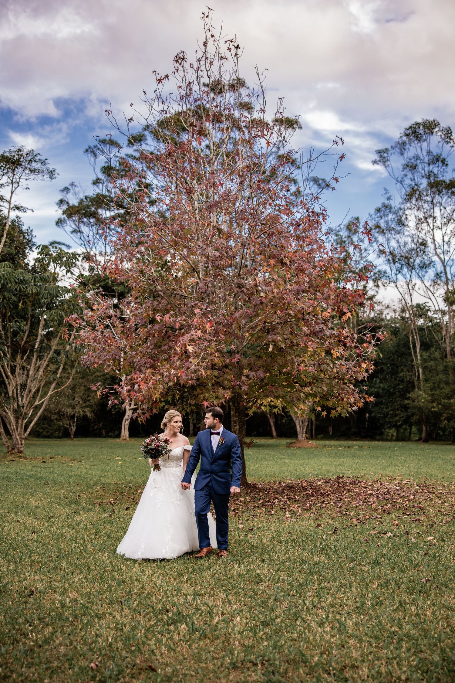 A couple takes wedding photos under a beautiful autumn tree with red leaves in a park setting.