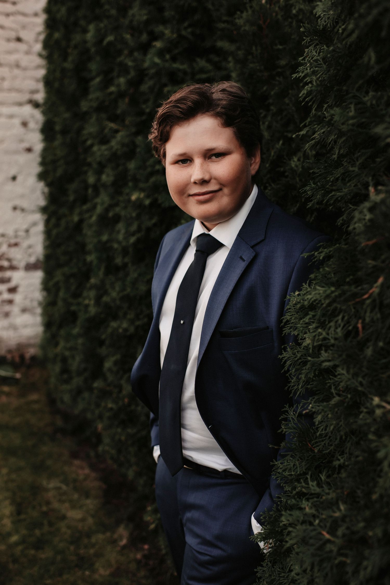 Portrait of a person in a navy blue suit and tie standing against an evergreen hedge.