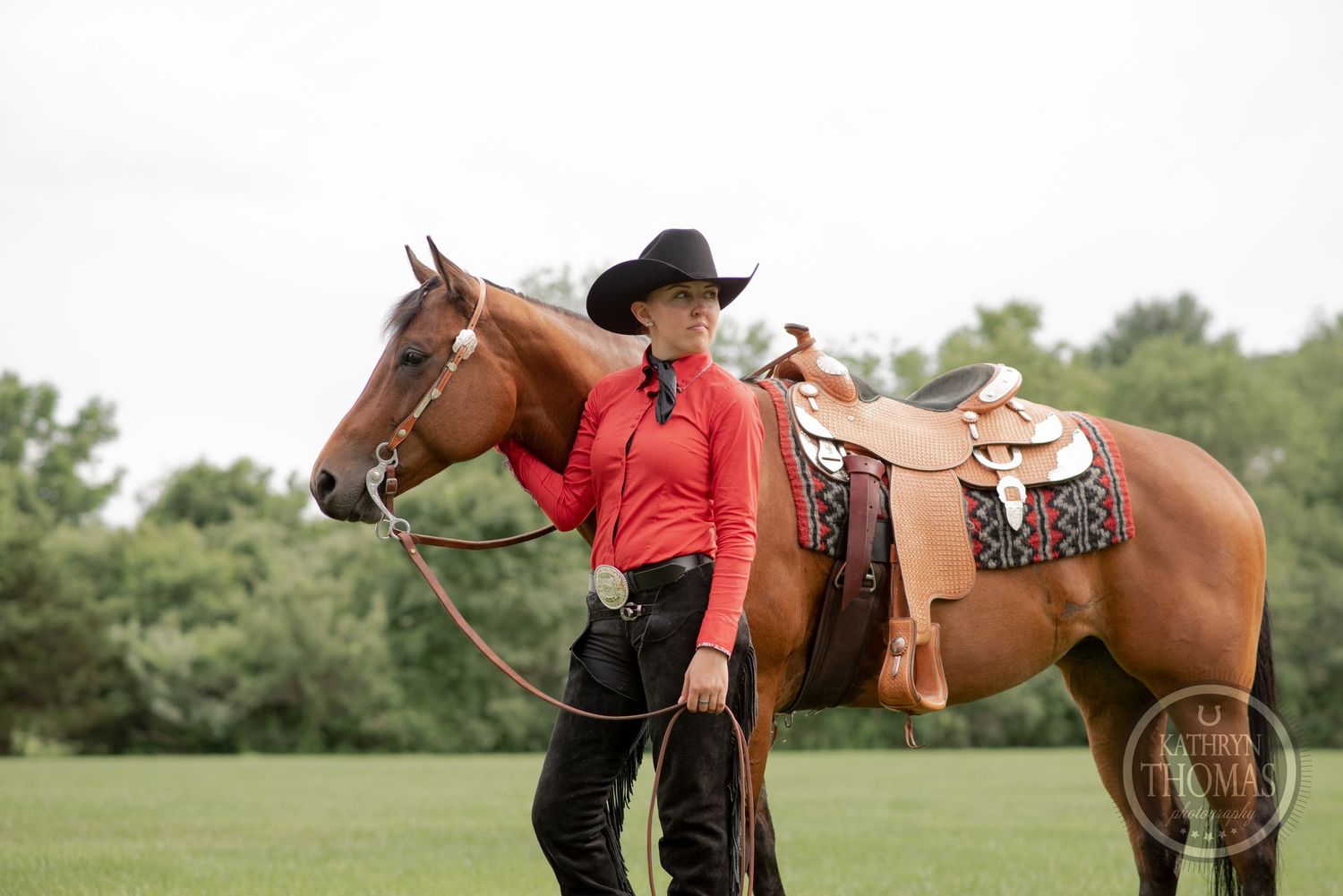 Brown horse with western saddle and handler in red shirt standing in green pasture.