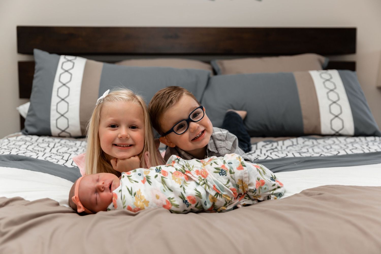 Bother an sister pose on the bed wth their new baby sister swaddled in front of them.